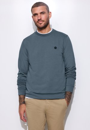 Mand med kort hår og skæg iført blå sweatshirt og beige bukser står mod en hvid baggrund med den ene hånd i lommen.