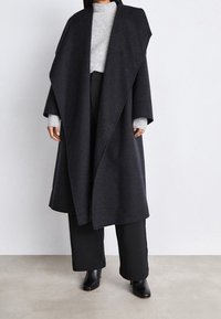 Langer, dunkelgrauer Wollmantel mit drapiertem Design, weiten Ärmeln und ohne sichtbare Verschlüsse, getragen über einem hellgrauen Pullover und schwarzen Hosen.