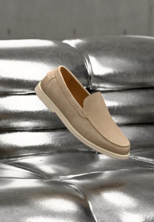 Chaussure beige en suède à enfiler avec semelle blanche flottant au-dessus d'une surface en cuir argenté matelassé et brillant.