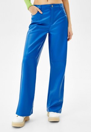 Kvinde iført lyseblå bukser med vidde ben og hvide kraftige sko, med den ene hånd i lommen og en kort neongrøn top.