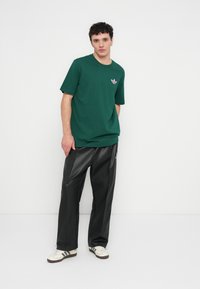 Zaļš kokvilnas T-krekls ar īsām piedurknēm un rozā Adidas logotipu uz krūtīm, kombinēts ar melnām mākslīgās ādas biksēm un baltām sporta kurpēm.