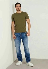 T-shirt verde oliva a maniche corte abbinato a jeans chiari strappati e sneakers bianche, su uno sfondo neutro.
