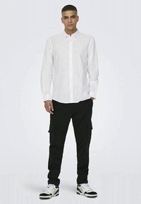 Camisa branca de botões com mangas longas, combinada com calças cargo pretas. A camisa possui um colarinho clássico e um tecido texturizado.
