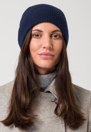 Bonnet tricoté bleu marine avec texture côtelée, porté sur un col roulé gris et un manteau gris clair, avec un détail de bouton près du col.