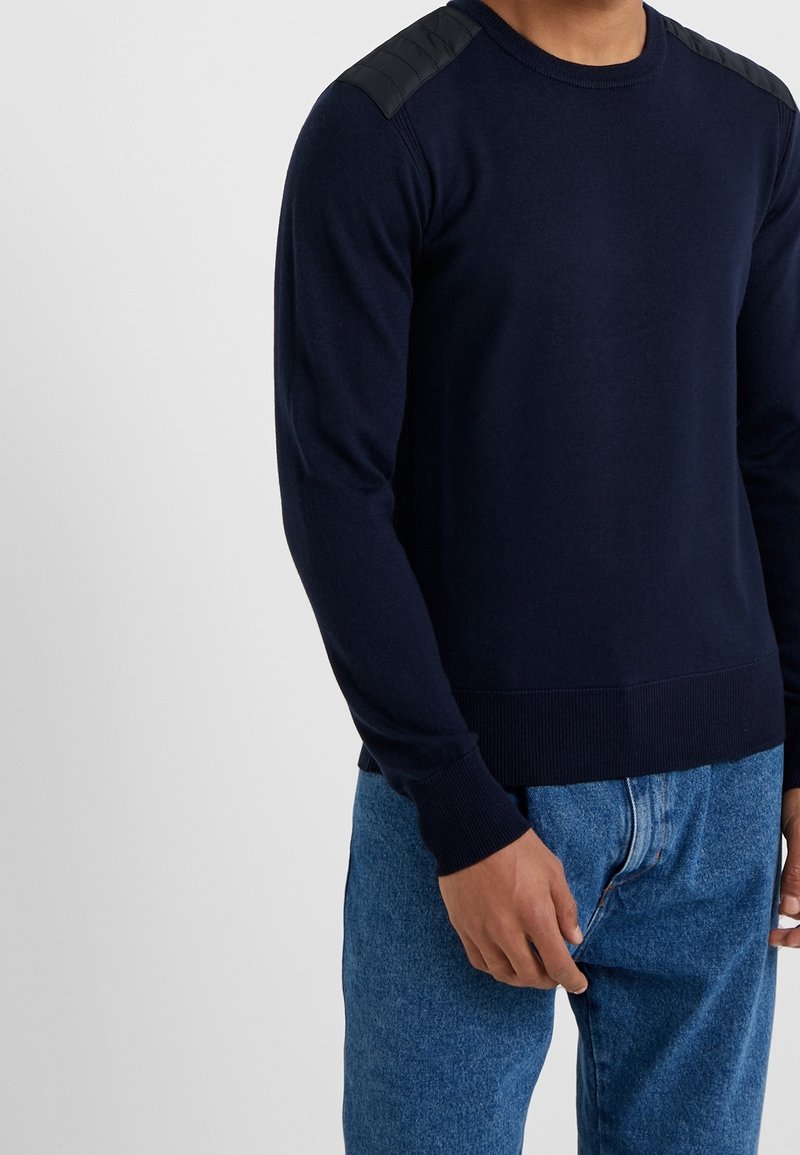 Homme portant un pull bleu marine à manches longues avec des renforts matelassés aux épaules et un jean bleu, debout devant un fond clair uni.