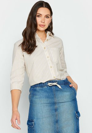 Camisa a rayas en beige claro y crema, con cuello y cierre de botones; combinada con una falda de mezclilla que tiene bolsillos y cintura con cordón.