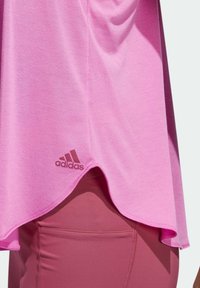 Gros plan sur un t-shirt rose Adidas avec ourlet arrondi porté sur un legging mauve avec poche latérale, montrant la texture du tissu et le détail du logo.