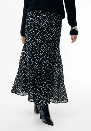 Kvinde iført sort plisseret midinederdel med hvide prikker, sort sweater, sølvarmbånd og sorte ankelstøvler med hæl, står på hvidt gulv.