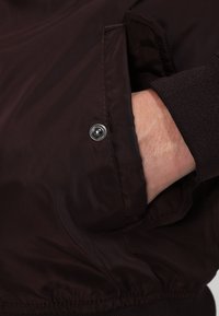 Giacca marrone leggera con una tasca con zip e dettaglio a bottone a pressione argentato. Tessuto liscio e cintura elasticizzata.