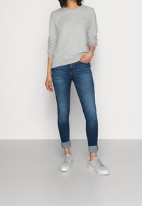 Felpa grigia con scollatura a girocollo, abbinata a jeans skinny blu scuro. Sneakers grigie con tomaia in tessuto texturizzato e suole bianche.