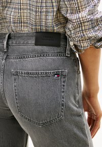 Γκρίζα τζιν παντελόνια από τζην ύφασμα με πίσω τσέπη που φέρει ένα μικρό μπλε, κόκκινο και λευκό λογότυπο. Μαύρη δερμάτινη ετικέτα με το "TOMMY HILFIGER" στη μέση της ζώνης.