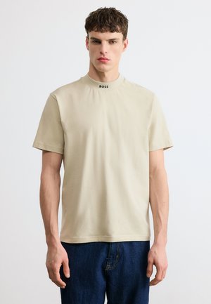 Νεαρός άνδρας που φοράει μπεζ κοντομάνικο πουκάμισο με το λογότυπο "BOSS" στον γιακά και σκούρο μπλε τζιν, στέκεται μπροστά σε έναν απλό φωτεινό φόντο.