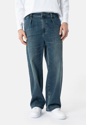 Uomo che indossa jeans di denim blu larghi, scarpe da ginnastica bianche e una camicia bianca a maniche lunghe, in piedi davanti a uno sfondo semplice.
