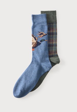 Deux paires de chaussettes, une bleue avec des motifs de canards volants, une vert olive avec un motif à carreaux, posées en chevauchement sur un fond uni.