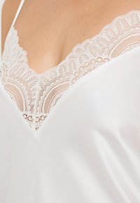 Camicetta in raso bianco con scollo a V adornato da un delicato bordo in pizzo bianco, che presenta intricati motivi floreali lungo il bordo.