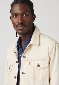 Beige spijkerjacket met bruine stiksel, drukknoopsluitingen en details op de borstzak. Gelaagd over een donkerblauwe spijkerblouse.