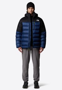 Granatowa i czarna kurtka puchowa z poziomymi przeszyciami, wysokim kołnierzem oraz kieszenią na zamek. Szare dresy i czarne buty sportowe noszone poniżej.