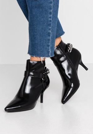 Bottes chelsea noires en cuir verni avec un bout pointu, un talon fin, une fermeture éclair sur le côté et un détail décoratif en chaîne argentée. Portées avec un jean à ourlet effiloché.