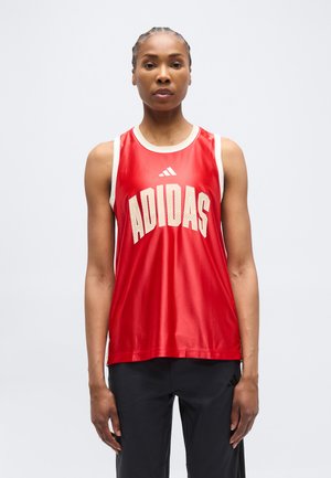 Femeie cu părul împletit poartă un top sport roșu fără mâneci Adidas și pantaloni sport negri, stând în picioare pe un fundal simplu, deschis la culoare.