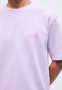 Άτομο που φοράει ανοιχτό μωβ μπλουζάκι με μικρό ροζ κείμενο "Lindbergh Beach & Resort Club" στο αριστερό στήθος, με ορατά κοντά μανίκια.