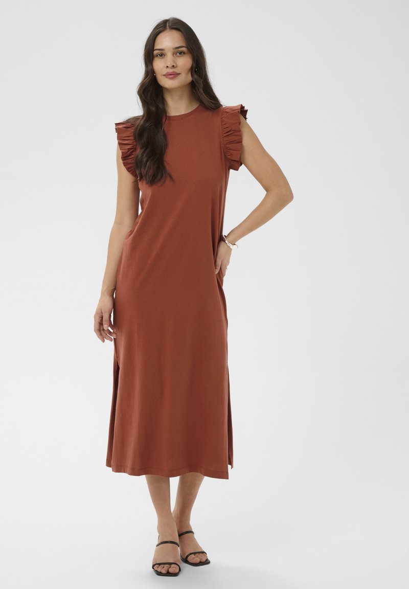 Femme aux longs cheveux foncés portant une robe midi sans manches couleur rouille avec des épaules à volants et des sandales noires à lanières, debout sur un fond blanc.