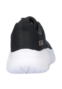 Zapatilla deportiva negra con una parte superior de malla texturizada, suela blanca contrastante y un pequeño logo metálico. Altura estándar del talón y bucle de agarre.