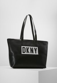 Černá kožená taška ve tvaru plochého pytle s dvojitými uchy a bílým kostkovaným logem DKNY na přední straně. Hladká textura.