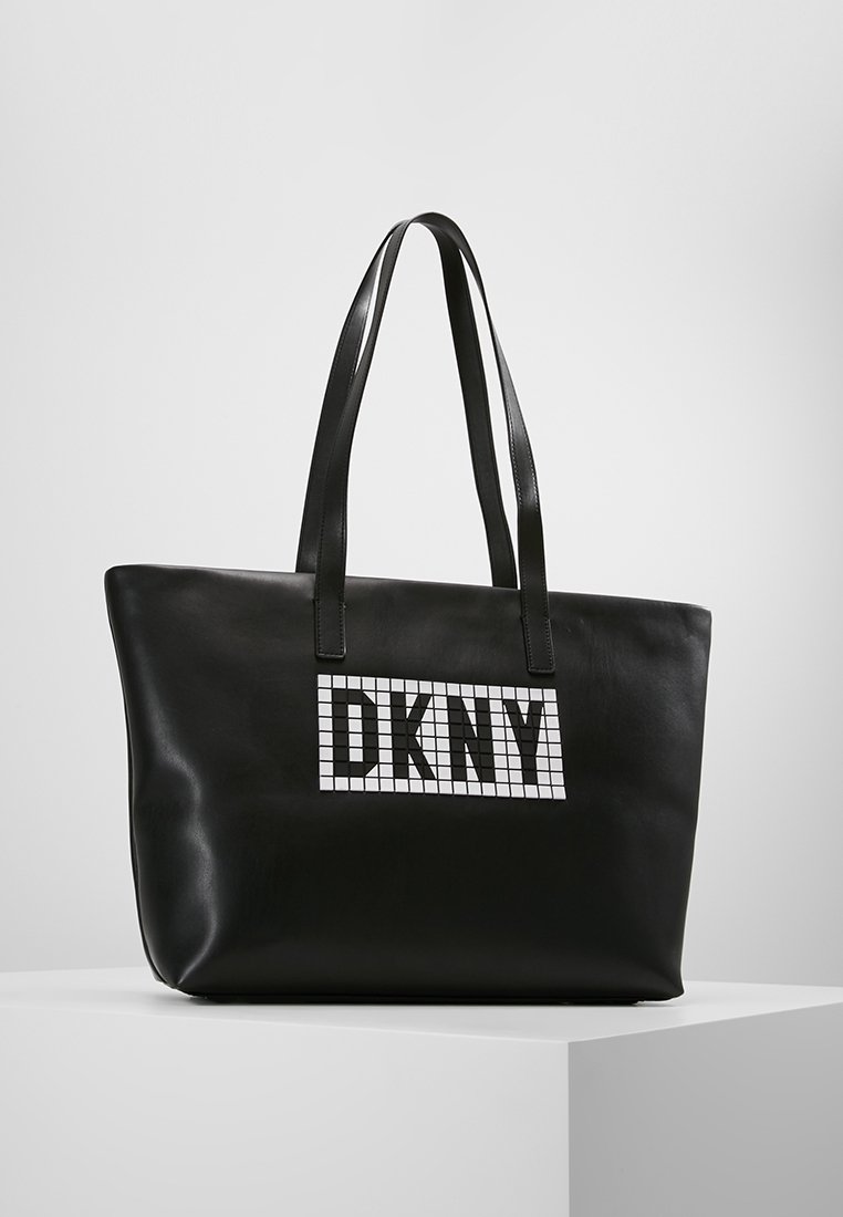 Černá kožená taška ve tvaru plochého pytle s dvojitými uchy a bílým kostkovaným logem DKNY na přední straně. Hladká textura.