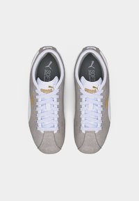 Ζευγάρι γκρι αθλητικά παπούτσια Puma με άνεση μαλακής σόλας, λευκά κορδόνια και χρυσό λογότυπο, απεικονιζόμενα από πάνω σε λευκό φόντο.