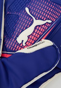 PUMA keepershandschoen met een blauwe latex buitenkant, textuur voor grip, roze accenten en een 'Thumb Wrap'-label. Close-up weergave van het ontwerp.