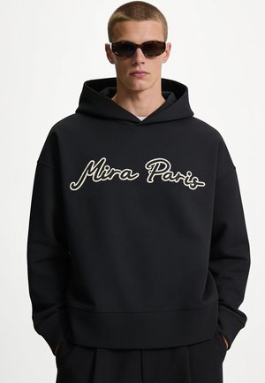 Mann trägt Schildpatt-Sonnenbrille und schwarzen Kapuzenpullover mit weißer, kursiver Aufschrift ""Mira Paris"" auf der Vorderseite.