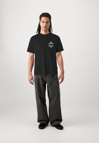 Musta puuvillapaita, jossa on värikäs geometrinen kuvio rinnassa, yhdistettynä rentoihin harmaisiin farkkuihin ja mustiin kenkiin.