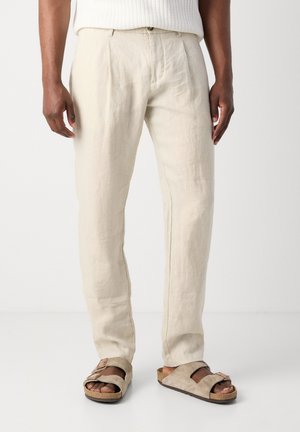 Man wearing cream linen pants and beige suede sandals against plain white background.