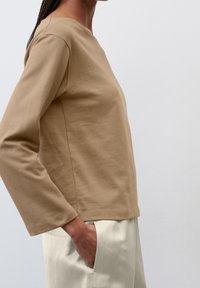 Beige langärmliges Baumwolloberteil mit rundem Halsausschnitt, Seitennähten und lockerer Passform, kombiniert mit hellen Hosen.