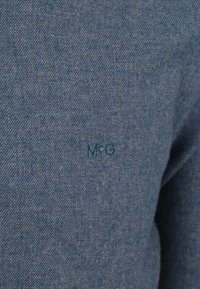 Blauwe stof met een gestructureerde weving, voorzien van een reliëf "MCG" logo in bijpassend blauw nabij de bovenste linkerschouder.