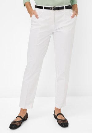 Woman wearing white tailored pants, black belt, black mesh flats, and a light green top with hands in pockets.