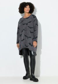Donna con capelli ricci che indossa una lunga tunica grigia con stampe di pipistrelli neri, abbinata a leggings neri lucidi e stivali neri con borchie.