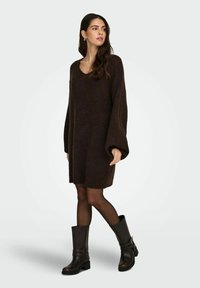 Brązowa dzianinowa sukienka z dekoltem w serek i oversize'owymi rękawami, zestawiona z czarnymi kozakami za kolano z paskami i masywnym obcasem.