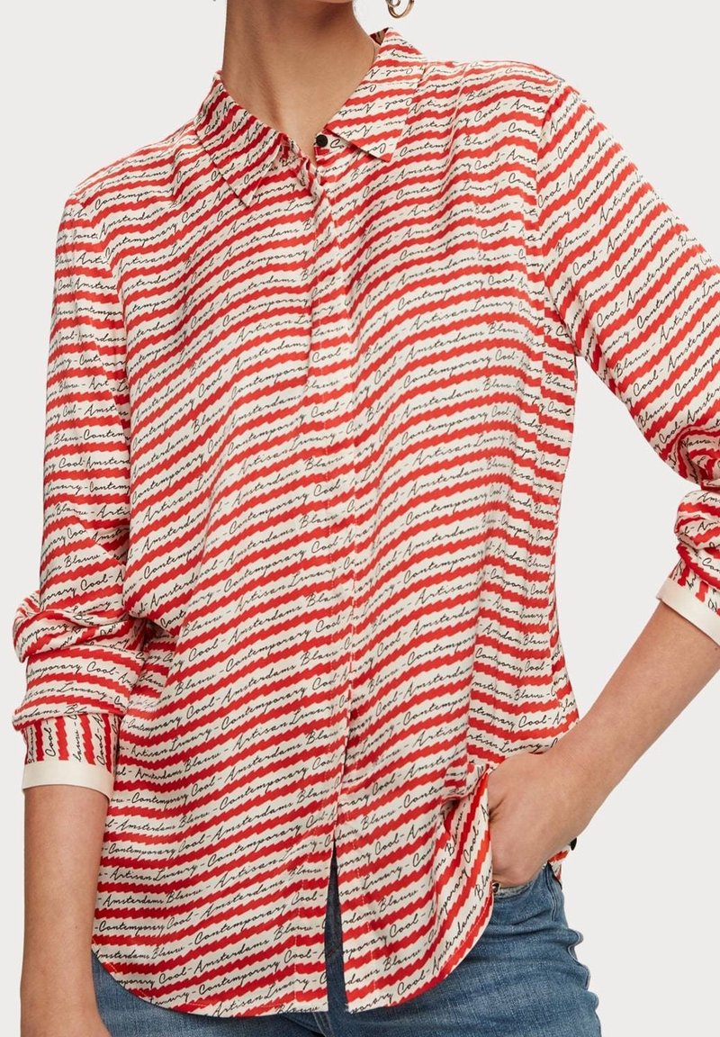 Femme portant une chemise à rayures rouges et blanches à boutons avec du texte noir en cursive et un jean bleu, main dans la poche, manches retroussées.
