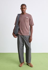 Tricou din bumbac maro cu buzunar și logo, asortat cu pantaloni largi gri și Adidași bej. Modelul poartă o geantă de umăr gri.