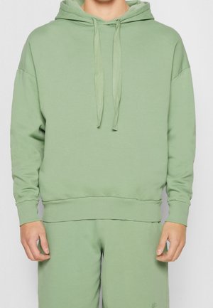Homme portant un sweat à capuche vert clair et un pantalon de jogging assorti, debout les bras détendus le long du corps sur fond blanc.