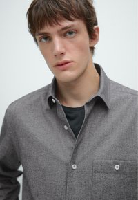 Grijze overhemd met een subtiele textuur, puntige kraag, borstzak en witte knopen; gelaagd over een zwart shirt daaronder.