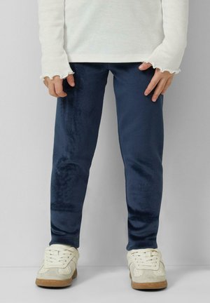 Child wearing navy blue velvet leggings, white long-sleeve top with ruffled cuffs, and white sneakers standing against a plain background.