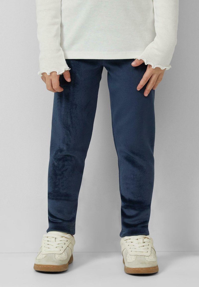 Child wearing navy blue velvet leggings, white long-sleeve top with ruffled cuffs, and white sneakers standing against a plain background.