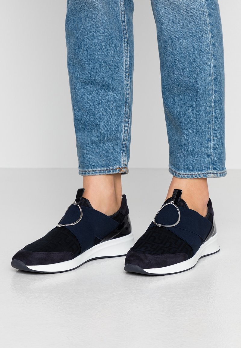 Personne portant un jean en denim bleu et des baskets à enfiler noires avec des semelles blanches et de grands détails de boucles en argent.