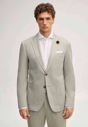 Junger Mann mit lockigem Haar, der einen hellgrauen strukturierten Anzug und ein weißes Hemd trägt und vor einem schlichten hellen Hintergrund posiert.