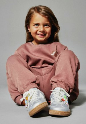 Uśmiechnięte dziecko siedzi skrzyżowanymi nogami, ubrany w fioletowy dres i srebrne trampki z kolorowymi koralikowymi zawieszkami na szarym tle.