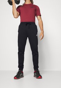 Röd kortärmad sporttröja, svarta avsmalnande joggingbyxor med dragsko och svarta sportskor med röda detaljer. Lyfter en hantel.