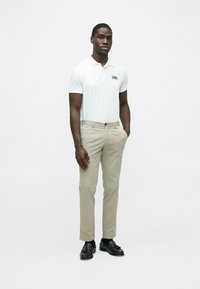 Weißes Poloshirt mit einem dezenten Logo, kombiniert mit hellbeigen Chinos und schwarzen Schuhen. Das Modell steht mit den Händen in den Taschen vor einem neutralen Hintergrund.