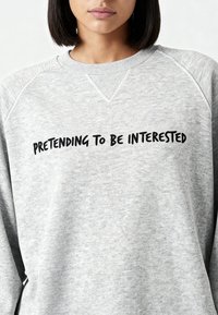 Sweatshirt gris à col rond et manches raglan, avec le texte noir « PRÉTENDRE ÊTRE INTÉRESSÉ » affiché en évidence sur le devant.